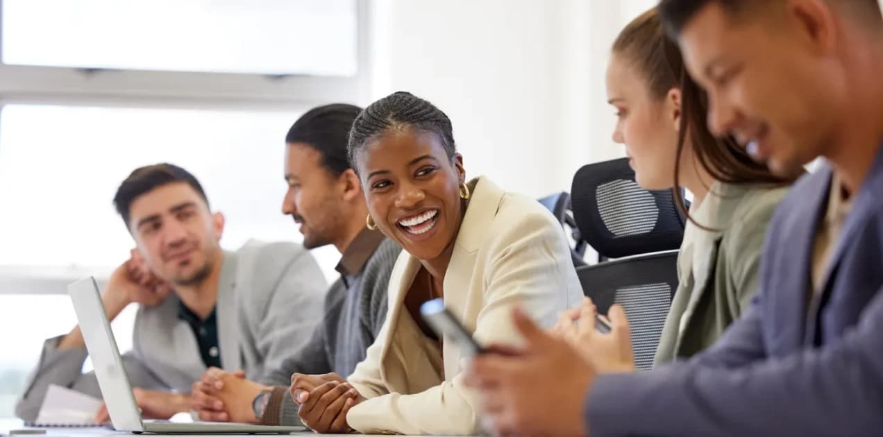 Colaboradores trabalhando na empresa. A imagem tem vários colaboradores trabalhando dentro da empresa, tanto homens quantos mulheres. Na mesa, tem notebooks e celulares.